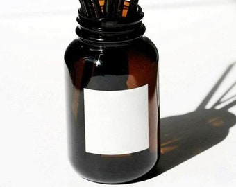 May include: A brown glass bottle with a white label and black sticks sticking out of the top. The bottle is sitting on a white surface.
