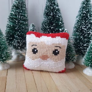 Pode incluir: Uma pequena almofada de ponto cruz branca e vermelha com um rosto de Papai Noel. A almofada está sentada em uma superfície de madeira na frente de três pequenas árvores de Natal artificiais verdes.