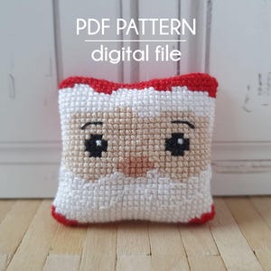 Pode incluir: Uma almofada de ponto cruz vermelha e branca com um rosto de Papai Noel. A almofada é pequena e tem uma barba branca e olhos pretos. O texto "PDF PATTERN digital file" está acima da almofada.