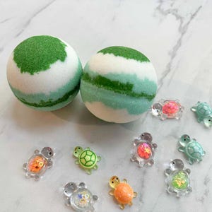 May include: Two round bath bombs with green and white layers, alongside several small, clear turtle-shaped embellishments with colorful interiors. The bath bombs have a textured, speckled appearance, and the turtles vary in color.