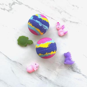 May include: Two colorful bath bombs with blue, pink, yellow, and purple swirls are surrounded by small, whimsical figurines. A green turtle, a pink pig, a purple dinosaur, and a pink unicorn are scattered around the bath bombs.