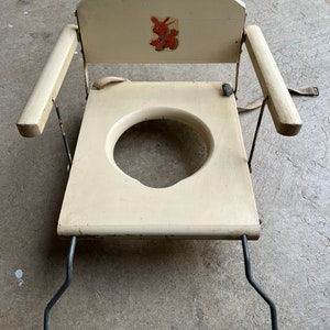 May include: A vintage white wooden potty chair with a bunny rabbit design on the back. The chair has two armrests and a metal frame.