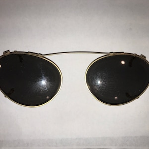 May include: A pair of gold-rimmed round sunglasses with black lenses. The sunglasses have a clip-on design.