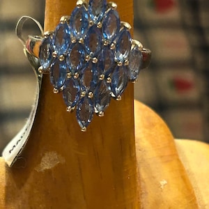 May include: A silver ring featuring a cluster of marquise-cut, light blue gemstones arranged in a diamond shape. The ring is displayed on a wooden surface, with a tag visible on the side.