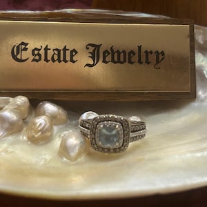 May include: A square-cut aquamarine ring with a halo of small diamonds, set in a silver band. The ring is displayed on a white shell with several pearls, next to a wooden sign that reads "Estate Jewelry".