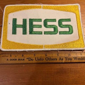 May include: Embroidered patch with the word "HESS" in green block letters on a white background, framed by a yellow border. A wooden ruler is below the patch.