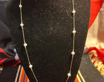 May include: A delicate gold necklace featuring small, evenly spaced pearls. The necklace is displayed on a black velvet bust, showcasing its elegant design. The chain is thin and the pearls are white.