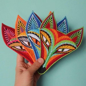 May include: Five colorful embroidered patches in the shape of a fox face. The patches are layered on top of each other, with the largest patch in the center. The patches are made of felt and have intricate details, including eyes, a nose, and a mouth.