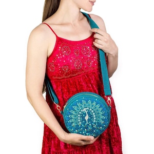 Peut inclure: Sac bandoulière rond bleu turquoise avec un motif de paon. Le sac a une bandoulière turquoise et une fermeture éclair dorée. La femme porte une robe rouge.