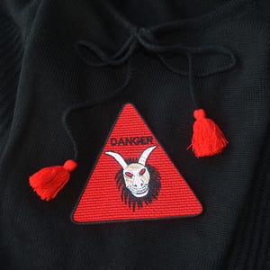 Könnte beinhalten: Schwarzes Kleidungsstück mit einem roten dreieckigen Aufnäher mit einem weißen Schädel mit Hörnern und dem Wort „DANGER“. Der Aufnäher ist mit roten Quasten verziert. Das Kleidungsstück hat einen Kordelzug.