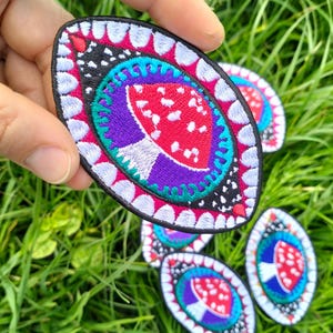 May include: Embroidered oval patch featuring a red mushroom with white spots, surrounded by a purple, teal, and black design. The patch has a white border with pink accents. Several patches are visible on a bed of green grass.