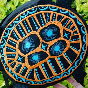May include: A black round bag with a blue and orange embroidered design. The design features a stylised skull with geometric patterns.