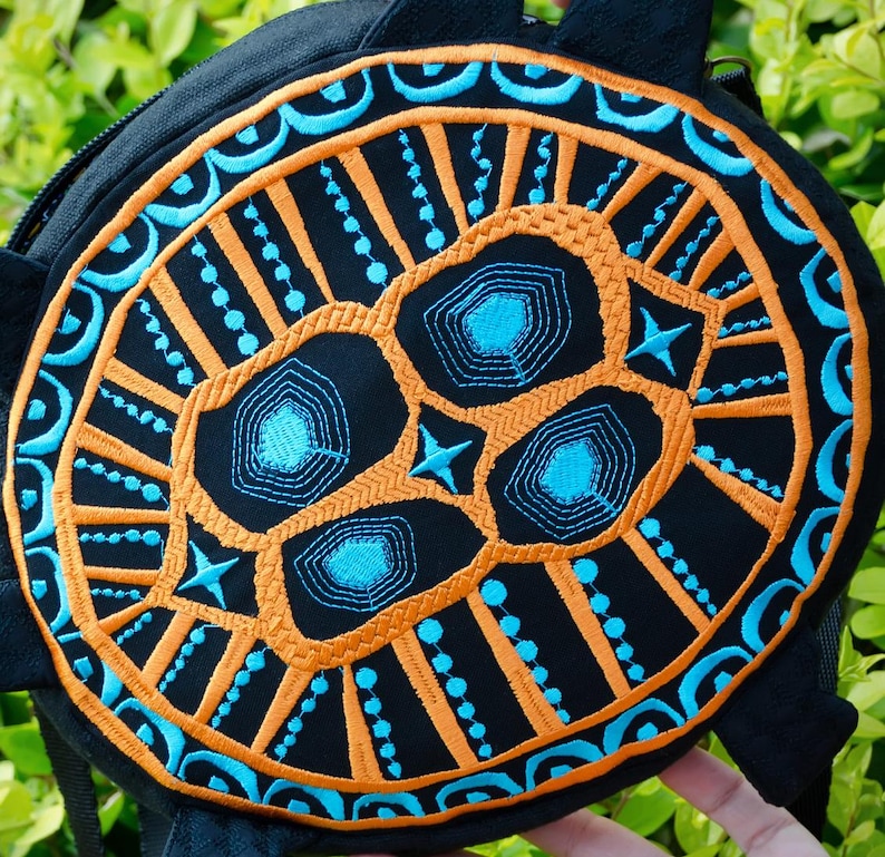 May include: A black round bag with a blue and orange embroidered design. The design features a stylised skull with geometric patterns.