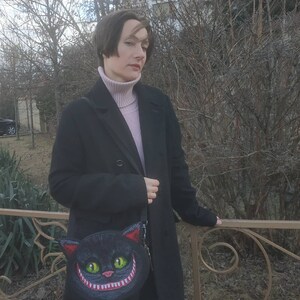May include: A woman wearing a black coat and a pink turtleneck sweater is holding a black purse with a grinning cat face design.
