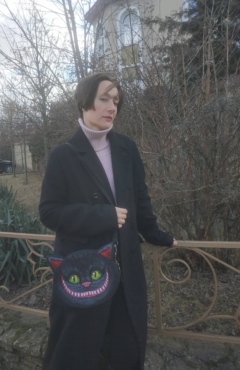 May include: A woman wearing a black coat and a pink turtleneck sweater is holding a black purse with a grinning cat face design.