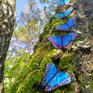 Puede incluir: Una fila de aplicaciones de mariposas azules y moradas están unidas a un tronco de árbol cubierto de musgo. Las mariposas están dispuestas en línea, ascendiendo por el tronco.