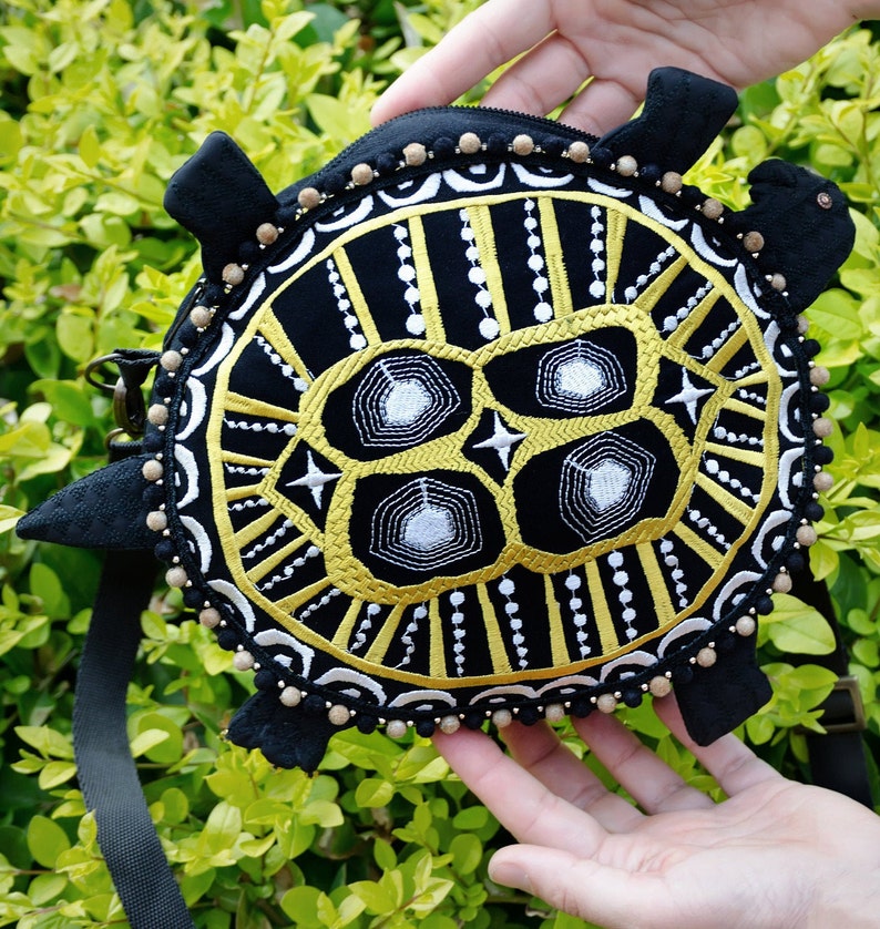 May include: A black turtle-shaped purse with a gold and white embroidered design. The purse has a black strap and a beaded trim.