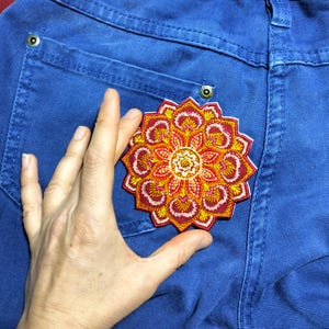 Peut inclure: Un patch brodé en forme de mandala rouge, orange et jaune avec un centre blanc. Le patch est repassé sur une poche en jean bleu.