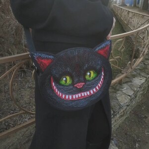 May include: A black crossbody bag with a grinning cat face design. The cat's eyes are green and the mouth is red with white teeth.