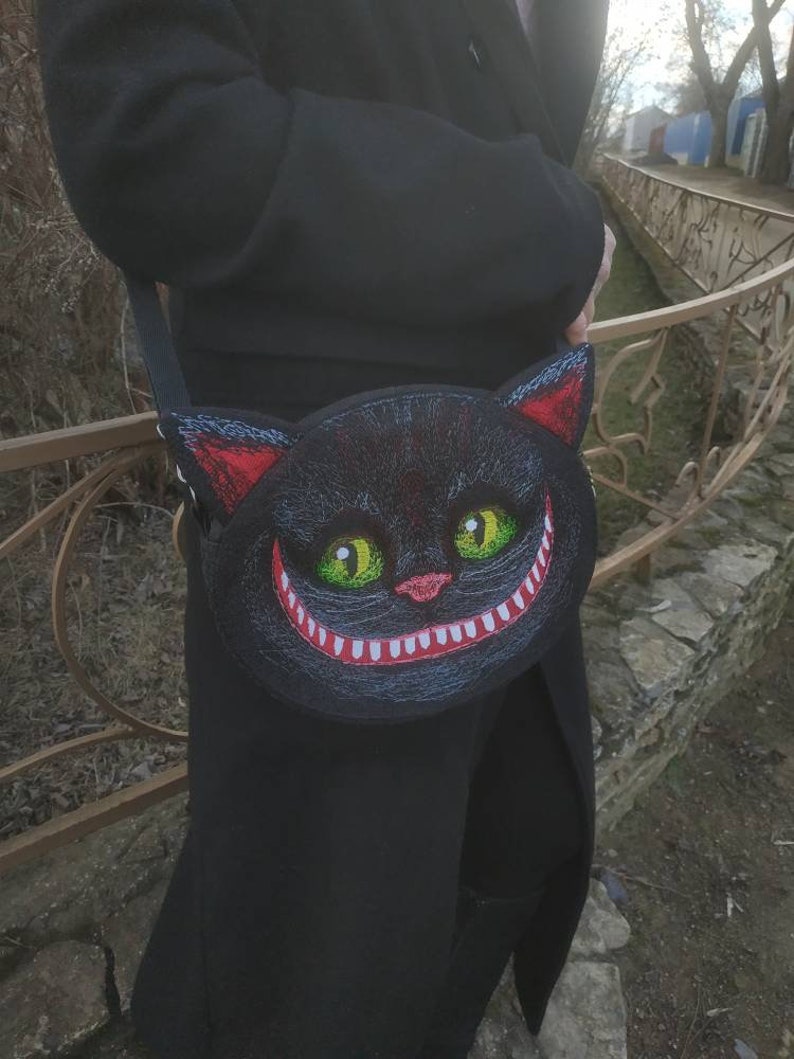 May include: A black crossbody bag with a grinning cat face design. The cat's eyes are green and the mouth is red with white teeth.