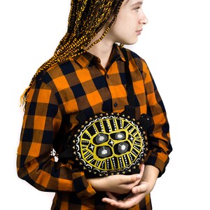 May include: A person with long black and yellow braids is wearing a plaid shirt and holding a black and yellow embroidered turtle cushion.