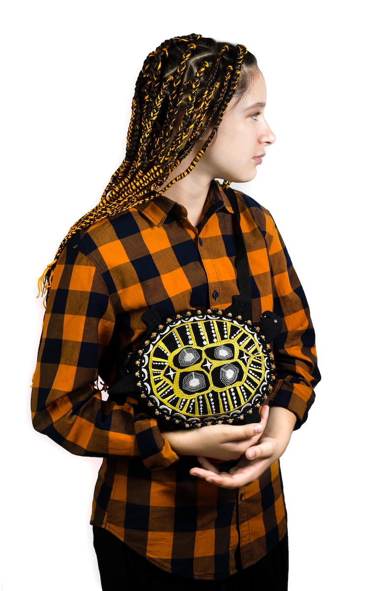 May include: A person with long black and yellow braids is wearing a plaid shirt and holding a black and yellow embroidered turtle cushion.
