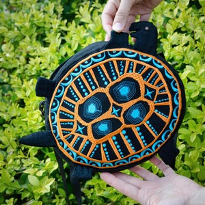 May include: A black and orange turtle-shaped bag with blue embroidery. The bag has a black strap and is being held in someone's hands.