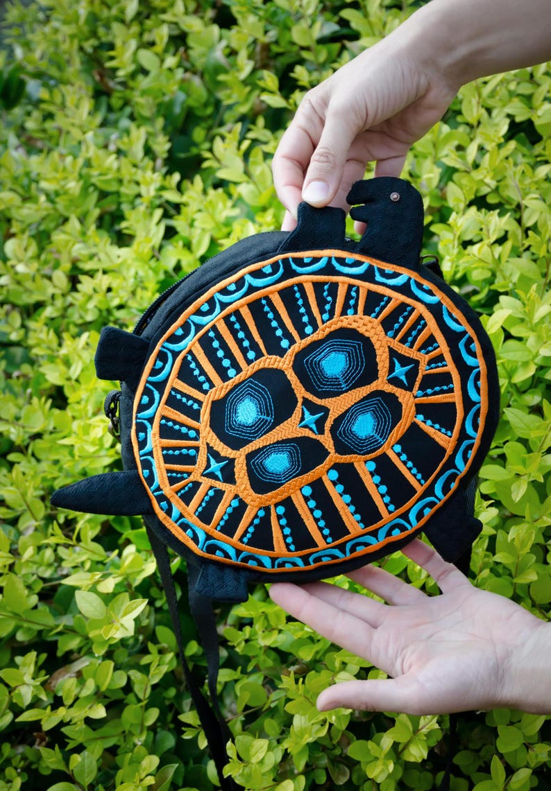 May include: A black and orange turtle-shaped bag with blue embroidery. The bag has a black strap and is being held in someone's hands.