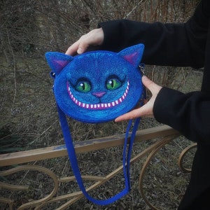 May include: A blue round purse with a white stitched grin and green eyes, resembling the Cheshire Cat from Alice in Wonderland. The purse has a blue strap.