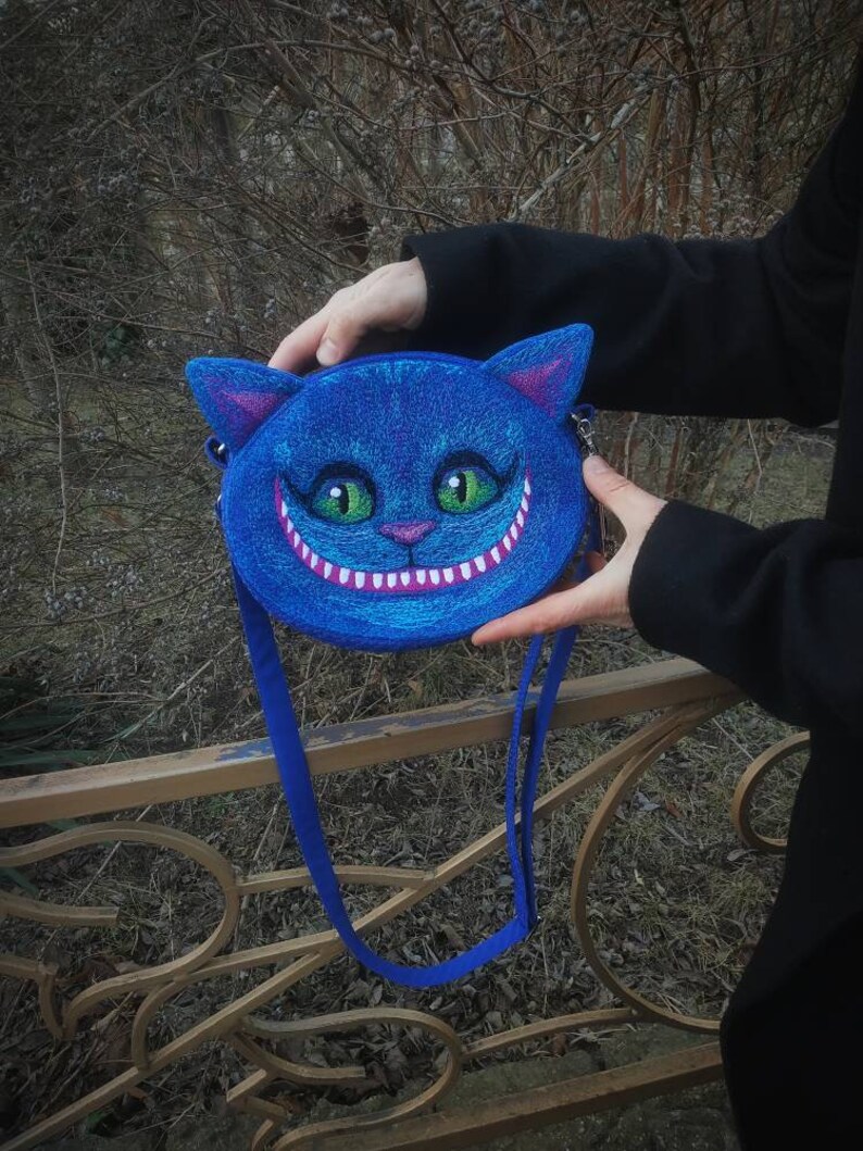 May include: A blue round purse with a white stitched grin and green eyes, resembling the Cheshire Cat from Alice in Wonderland. The purse has a blue strap.