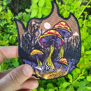 May include: Embroidered patch featuring a whimsical mushroom scene. The patch showcases colorful mushrooms with red, yellow, and purple caps, set against a backdrop of a forest and a full moon. The patch has a brown border.