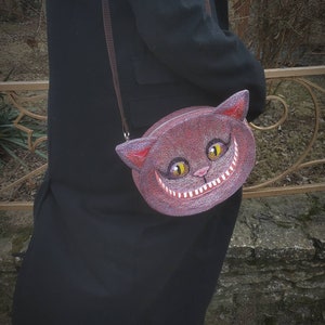 May include: A brown felt purse shaped like a cat's face with a wide grin and yellow eyes. The purse has a brown strap and is worn over the shoulder.