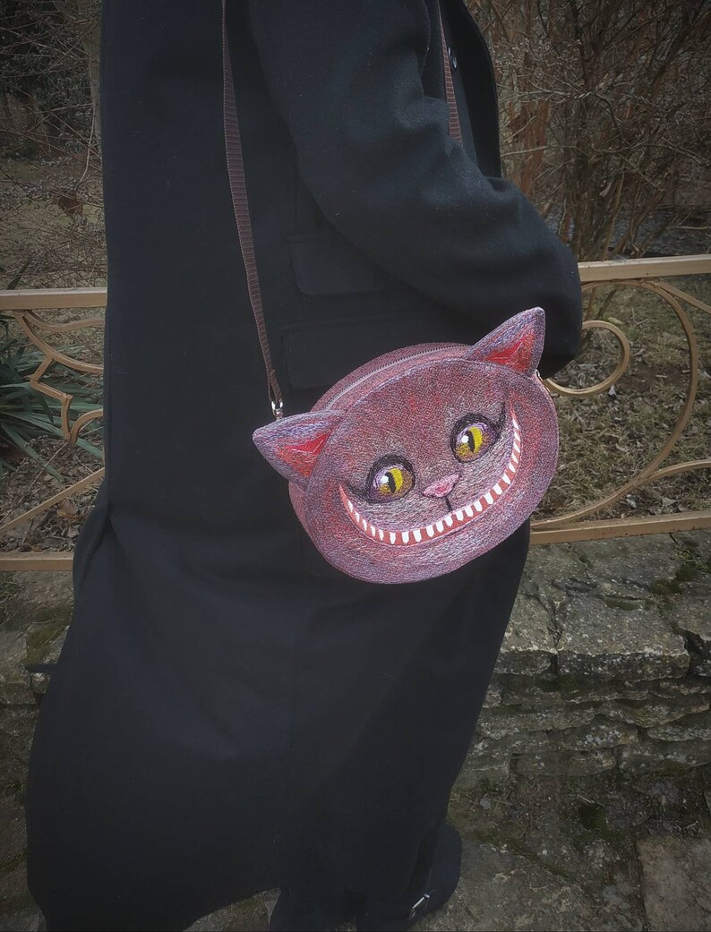May include: A brown felt purse shaped like a cat's face with a wide grin and yellow eyes. The purse has a brown strap and is worn over the shoulder.