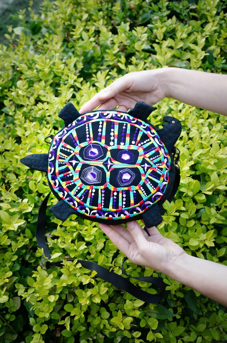 May include: A black turtle-shaped purse with a colourful embroidered design. The embroidery features a geometric pattern with purple, red, yellow, green, and blue accents.