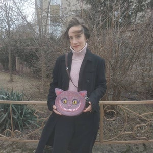 May include: A woman wearing a black coat and a pink turtleneck sweater is holding a purple and brown purse shaped like a cat's face with a wide grin. The purse has yellow eyes and white teeth.