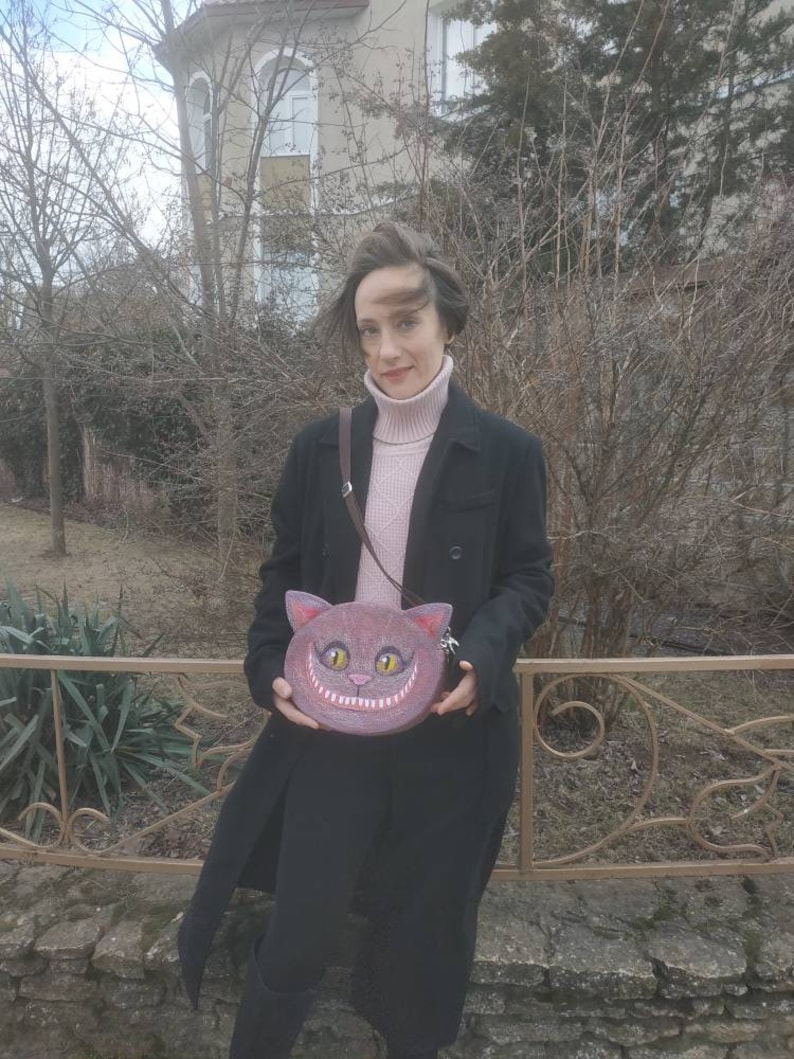 May include: A woman wearing a black coat and a pink turtleneck sweater is holding a purple and brown purse shaped like a cat's face with a wide grin. The purse has yellow eyes and white teeth.