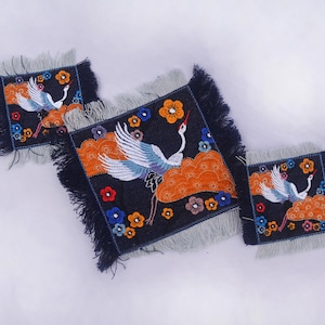 May include: Set of three square coasters with a dark blue background and a crane design. The crane is white and blue, with orange clouds and colorful flowers. The edges have a fringed trim.
