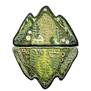 Puede incluir: Un parche bordado que representa una escena de montaña con árboles, un ciervo y un pájaro. El parche está dividido en dos secciones, la parte superior muestra la montaña y la parte inferior muestra el reflejo de la montaña en un lago.