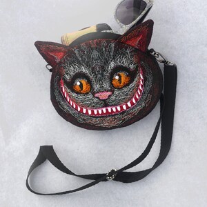 May include: A black and red embroidered cat face purse with a wide grin and large orange eyes. The purse has a black adjustable strap and a pair of sunglasses are tucked into the top of the purse.