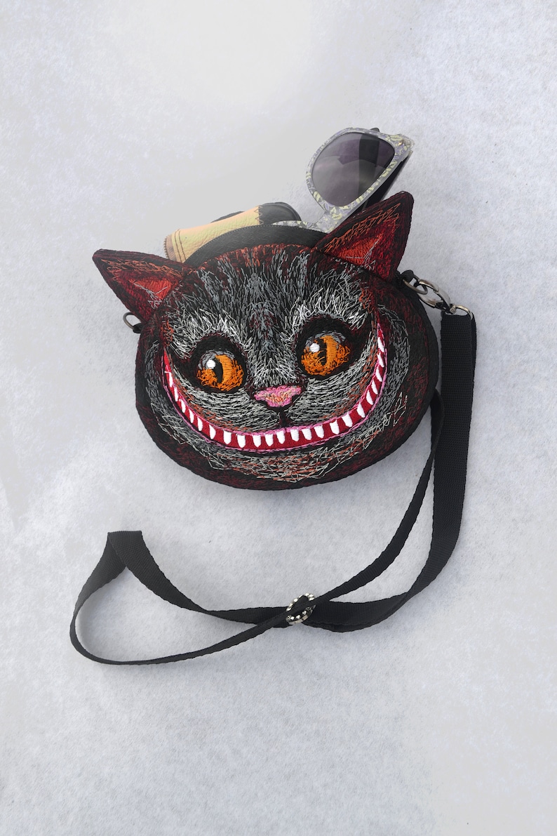 May include: A black and red embroidered cat face purse with a wide grin and large orange eyes. The purse has a black adjustable strap and a pair of sunglasses are tucked into the top of the purse.