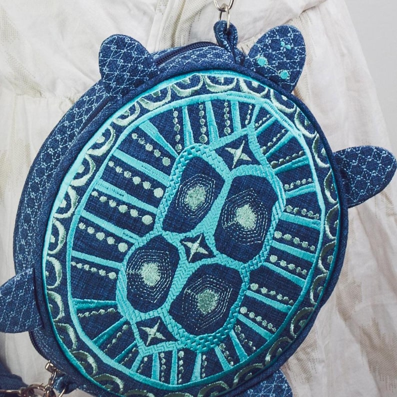 May include: A blue and teal round purse shaped like a turtle with a geometric pattern. The purse has a zipper closure and a strap.