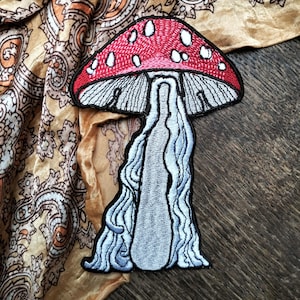 Op de afbeelding: Een geborduurde paddenstoelpatch met een rode en witte dop en een witte steel. De patch is op een bruine stoffen achtergrond.