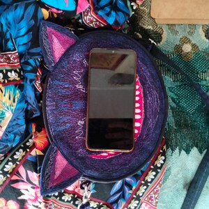 May include: A round, purple and blue cat-shaped purse with a pink embroidered nose and ears. A black smartphone is inside the purse.