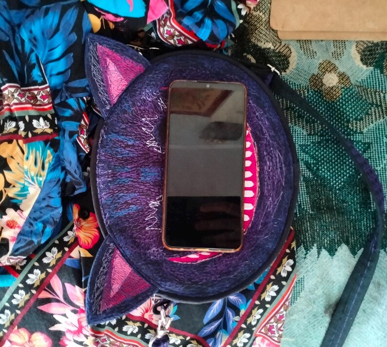 May include: A round, purple and blue cat-shaped purse with a pink embroidered nose and ears. A black smartphone is inside the purse.