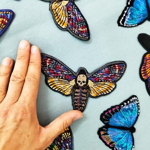 Könnte beinhalten: Eine Sammlung bunter, gestickter Bügeleisen-Patches mit verschiedenen Schmetterlings- und Mottenmotiven. Die Patches zeichnen sich durch aufwendige Details und leuchtende Farben aus, darunter Blau, Lila, Gelb und Rot.