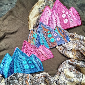 May include: Four embroidered iron-on patches featuring a mountain range design. The patches are in shades of pink, blue, and brown. The patches are arranged in a stack, with the largest patch at the bottom and the smallest patch at the top.