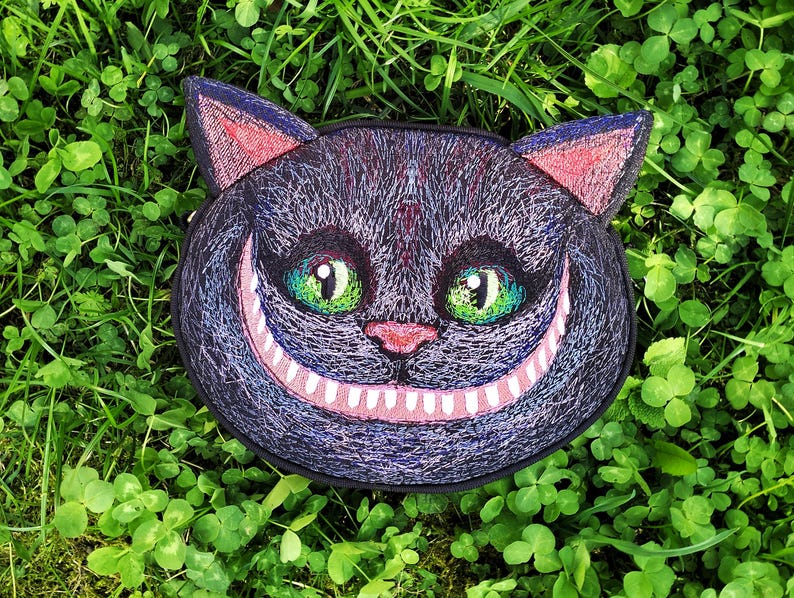 May include: A black and purple embroidered patch of a Cheshire Cat with a wide, toothy grin and green eyes. The cat is set against a green background.