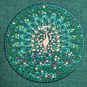 Puede incluir: Diseño circular bordado con un pavo real con las plumas de la cola extendidas. El pavo real es de color rosa claro, rodeado de detalles turquesa, verdes y rosas. El fondo es de tela verde oscuro, y el diseño está centrado.