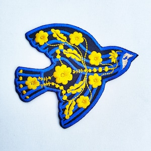 Puede incluir: Parche bordado azul y amarillo con detalles florales de un pájaro. El pájaro está volando con las alas extendidas. Las flores son amarillas y tienen un diseño simple.