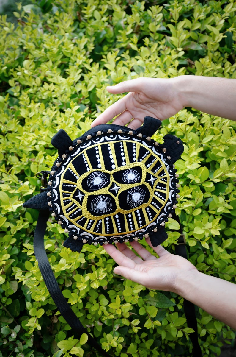 May include: A black and gold embroidered turtle-shaped purse with a black strap. The purse has a geometric design with white accents.